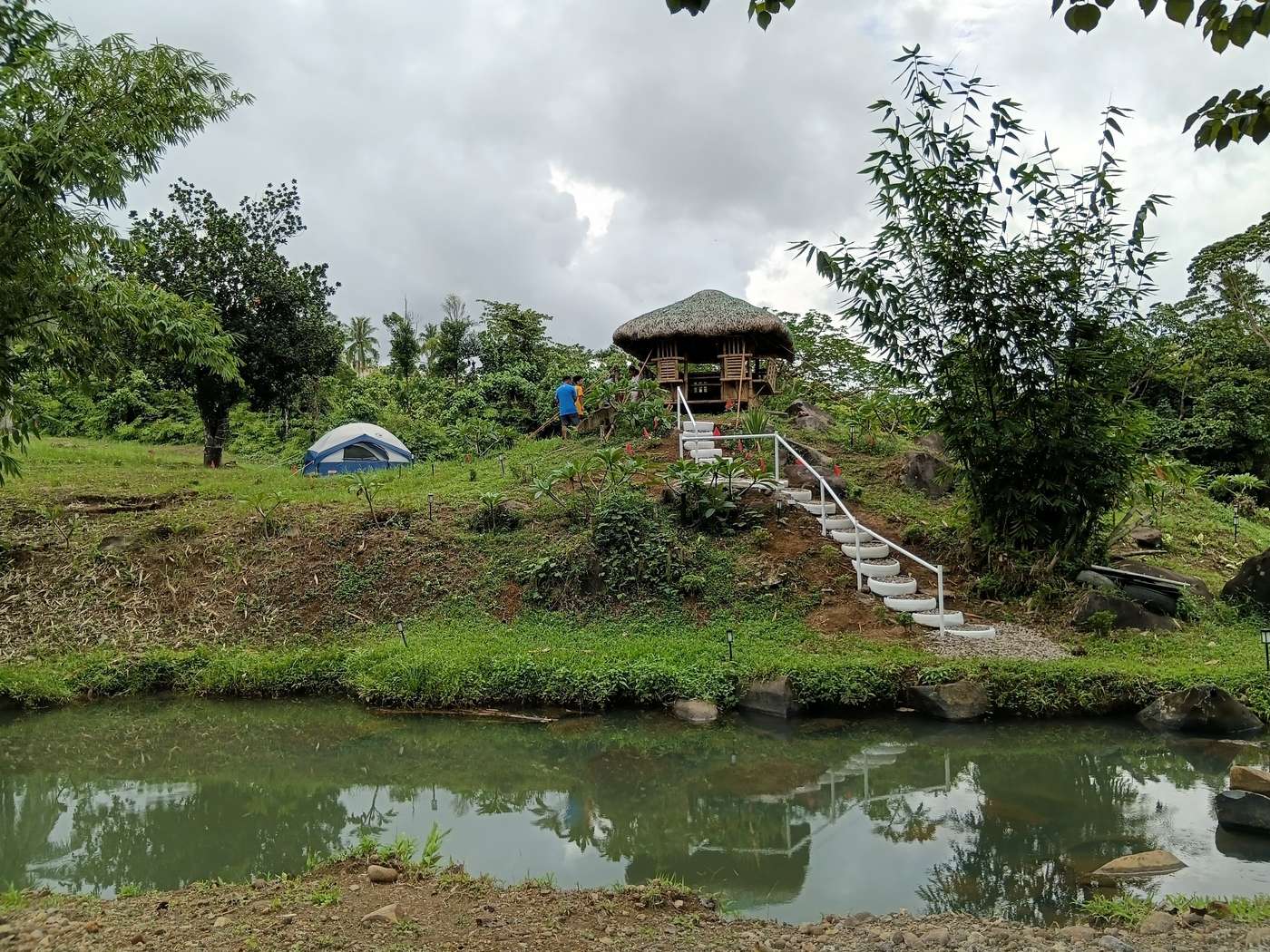 View across the water to the thatched kubo on the hill, with a tent on the lawn and a few people standing near the kubo